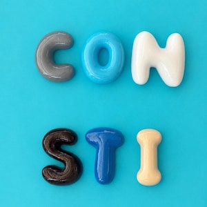 Könnte beinhalten: Ein Set aus sechs glänzenden, dreidimensionalen Buchstaben, die in zwei Reihen auf einem hellen Cyan-Hintergrund angeordnet sind. Die obere Reihe bildet CON in Grau, Cyan und Weiß. Die untere Reihe bildet STI in Dunkelbraun, Königsblau und Beige.