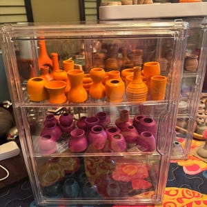 Könnte beinhalten: Eine transparente Kunststoff-Vitrine gefüllt mit kleinen, handgefertigten Keramikvasen. Die Vasen sind in Orange- und Rosatönen gehalten und weisen verschiedene Formen und Texturen auf. Die Vitrine hat drei Regale, und die Vasen sind ordentlich angeordnet.