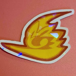 May include: A yellow and orange sticker with a flame-like design and a white border. The sticker has a holographic effect, creating a shimmering appearance. The sticker is on a pink background.