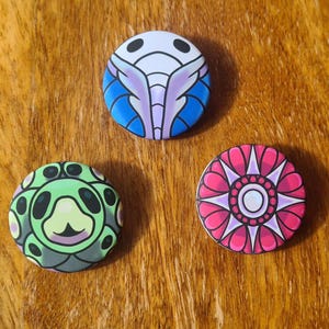 Puede incluir: Tres insignias redondas con diseños coloridos. Una presenta un insecto estilizado azul y morado, otra una criatura verde y la tercera un patrón floral rosa. Las insignias están sobre una superficie de madera.