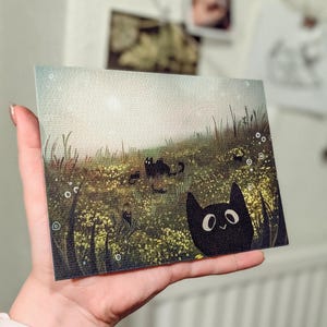 May include: A rectangular art print featuring a whimsical illustration of black cats in a field of yellow flowers and tall green grass. One cat peeks from the right corner. The background has a soft, light-filled sky.