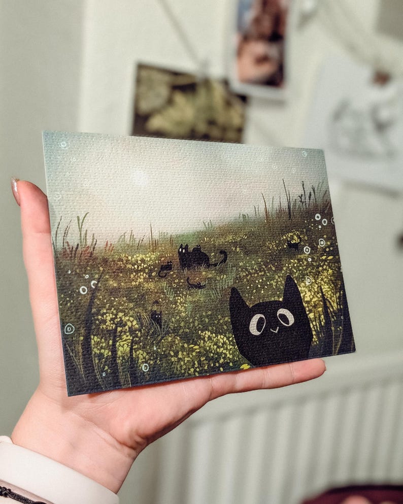 May include: A rectangular art print featuring a whimsical illustration of black cats in a field of yellow flowers and tall green grass. One cat peeks from the right corner. The background has a soft, light-filled sky.