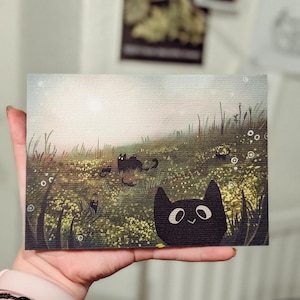 May include: A whimsical art print featuring a field of yellow flowers and several black cat illustrations. The artwork has a soft, dreamy quality with a smiling black cat in the foreground. The print is held in a hand.