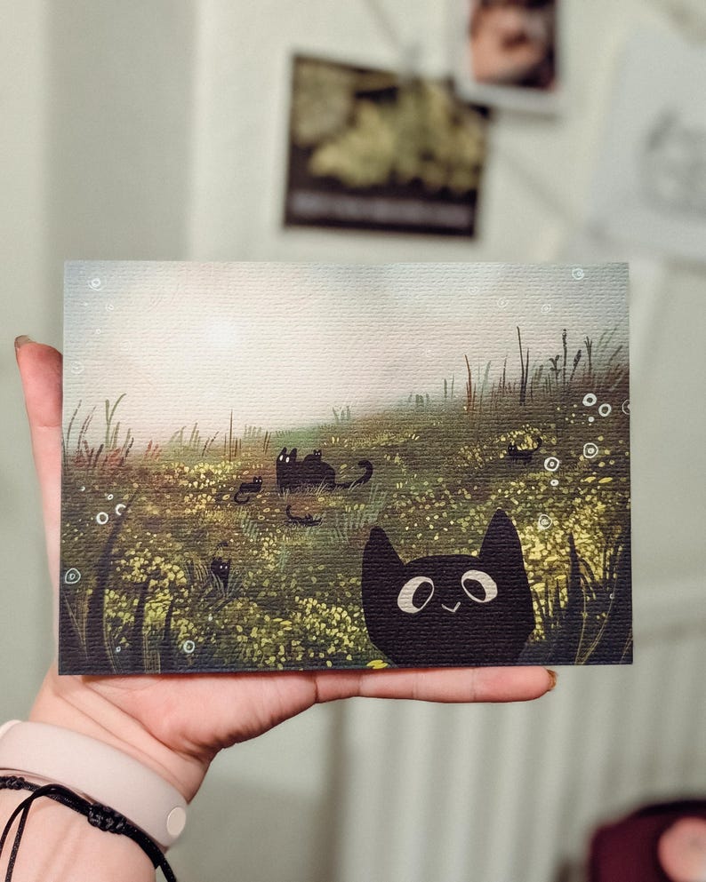 May include: A whimsical art print featuring a field of yellow flowers and several black cat illustrations. The artwork has a soft, dreamy quality with a smiling black cat in the foreground. The print is held in a hand.