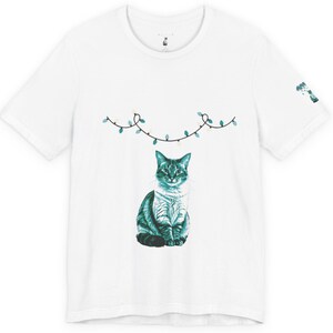 May include: White t-shirt featuring a teal illustration of a cat with a string of lights above it. The cat is sitting upright, with detailed fur and facial features. A small graphic is on the sleeve. The shirt is made of a soft material.