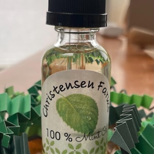 May include: A clear glass bottle of mint oil with a black dropper top. The label reads "Christensen Farms 100% Mint Oil" with a green leaf illustration. Green paper decorations surround the bottle.