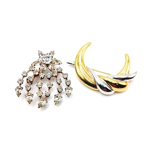 May include: Two vintage brooches. One is a starburst design with clear rhinestones and a copper-toned metal. The other is a gold and silver-toned abstract design. Both brooches are on a white background.