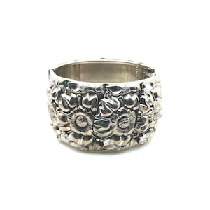 May include: A wide silver-toned hinged cuff bracelet featuring a detailed relief pattern of sunflowers and leaves. The metal surface has a polished finish with darkened recessed areas to highlight the floral design, and a secure metal hinge closure.
