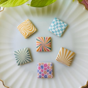 Könnte beinhalten: Sechs quadratische dekorative Magnete mit farbenfrohen Retro-Designs. Die Magnete weisen Muster wie Wirbel, Schachbrettmuster und Blumendrucke in Gelb-, Blau-, Rosa- und Orangetönen auf. Sie sind auf einem weißen, wellenförmigen Teller angeordnet.