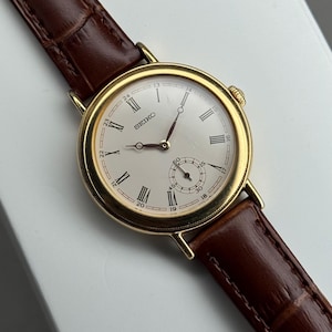 May include: A gold-toned Seiko wristwatch with a cream-colored face and Roman numeral hour markers. The watch features a small seconds dial and is paired with a brown leather strap.