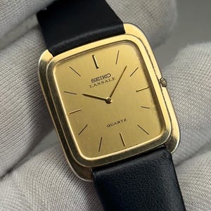 May include: A gold-toned Seiko Lassale quartz watch with a rectangular face and black leather band. The watch face displays the brand name and the word "QUARTZ". The watch has a classic, minimalist design.