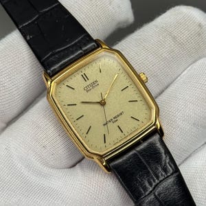 May include: A gold-plated Citizen Eco-Drive watch with a rectangular face and a black leather strap. The watch face is a light gold colour with black hour markers and hands. The watch reads "WATER RESIST 5 bar".