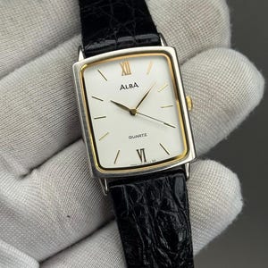 May include: Alba quartz watch with a rectangular face, white dial, gold hands and accents, and a black leather strap. The watch face displays the brand name and the word "QUARTZ". The watch is held against a white fabric background.