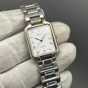 Puede incluir: Reloj Seiko Lucent rectangular con esfera blanca, agujas doradas y números romanos dorados. La correa es de plata y oro. El reloj se muestra sobre un fondo neutro.