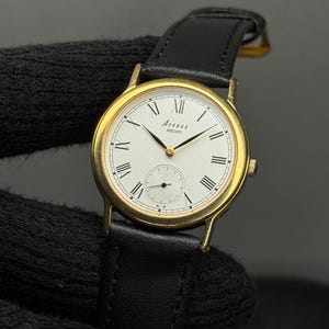 Puede incluir: Un reloj Avenue Seiko dorado con esfera blanca, números romanos negros y un pequeño dial de segundos. El reloj tiene una correa de cuero negro. El reloj es de diseño clásico.