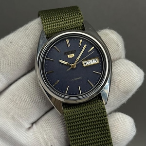 Puede incluir: Un reloj de pulsera plateado con esfera azul marino y agujas e indicadores dorados. El reloj tiene una pantalla de día y fecha y está colocado en una correa de nailon verde. La esfera del reloj indica "5" y "Automatic".