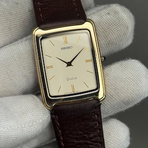 May include: A rectangular gold-toned Seiko Dolce watch with a cream-coloured face and a brown leather strap. The watch face features gold-coloured hands and markers, with the word "Dolce" printed below the centre. The strap is a dark brown colour.