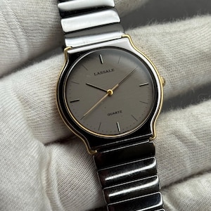 May include: A Lassale quartz wristwatch with a gray face and gold-tone accents. The watch features a silver-tone band and a minimalist design. The word "QUARTZ" is printed on the watch face.