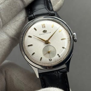 May include: A vintage Orient Star wristwatch with a silver-tone case and a white dial. The watch features gold-tone hands and markers, a small seconds subdial, and a black leather strap. The watch is held by a gloved hand.