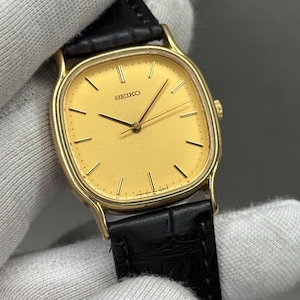 May include: A gold-coloured Seiko watch with a rectangular face and a black leather strap. The watch face is a light gold colour with gold hands and markers. A classic watch design.
