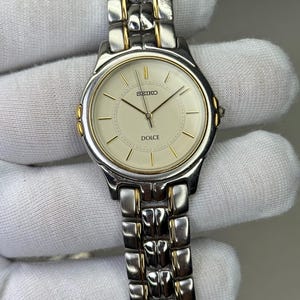 Pode incluir: Um relógio de pulso Seiko Dolce com uma pulseira de metal prateado e dourado. O mostrador do relógio é creme com ponteiros e marcadores de hora dourados. O relógio tem as palavras "SEIKO" e "DOLCE" impressas.