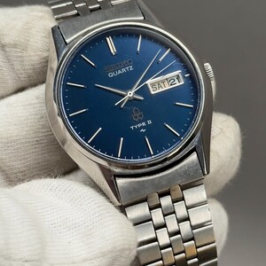 Op de afbeelding: Een zilveren Seiko Quartz Type II horloge met een blauwe wijzerplaat. Het horloge heeft zilveren wijzers en markeringen, een dag-datumweergave en een roestvrijstalen armband. De wijzerplaat toont de dag als "ZA 21".