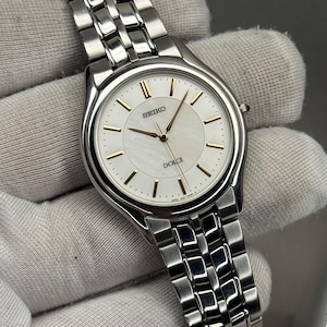 Op de afbeelding: Een zilverkleurig Seiko Dolce polshorloge met een ronde wijzerplaat van wit parelmoer, voorzien van goudkleurige uurmarkeringen en wijzers. Het horloge heeft een gepolijste roestvrijstalen schakelarmband en een kroon aan de rechterkant.