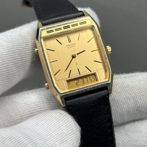 Peut inclure: Une montre Seiko Quartz dorée avec un cadran rectangulaire et un bracelet en cuir noir. Le cadran affiche des aiguilles analogiques et un affichage numérique de l'heure. La montre est de style vintage.