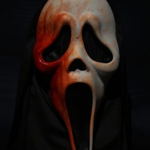 Puede incluir: Una máscara blanca de Scream con pintura roja que simula sangre en el lado izquierdo y salpicaduras por toda la cara. La máscara tiene grandes aberturas para los ojos y una boca ancha y abierta.