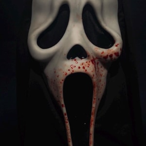 Puede incluir: Una máscara blanca de Scream con aberturas negras para los ojos y la boca. Salpicaduras y vetas rojas, similares a la sangre, son visibles en la máscara, añadiendo un elemento de terror. La máscara está sobre un fondo oscuro.