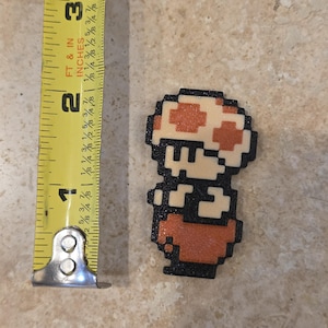 Puede incluir: Una figura pixelada de un personaje de hongo de un videojuego, de aproximadamente 5 cm de altura. El personaje tiene un sombrero de hongo blanco y naranja, un contorno negro y un cuerpo rojo-naranja. La imagen est&aacute; tomada desde arriba.