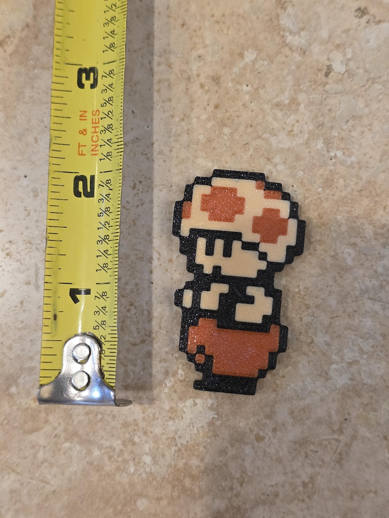 Puede incluir: Una figura pixelada de un personaje de hongo de un videojuego, de aproximadamente 5 cm de altura. El personaje tiene un sombrero de hongo blanco y naranja, un contorno negro y un cuerpo rojo-naranja. La imagen est&aacute; tomada desde arriba.