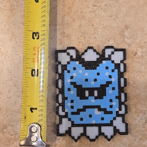 Puede incluir: Un dise&ntilde;o de personaje pixelado, azul y negro, de aproximadamente 6,35 cm de altura, con un borde blanco. El personaje tiene una expresi&oacute;n severa. El dise&ntilde;o es probablemente una referencia a un videojuego.
