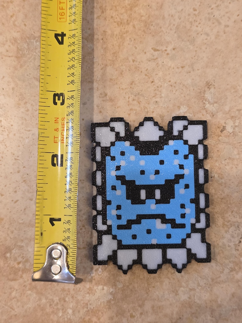 Puede incluir: Un dise&ntilde;o de personaje pixelado, azul y negro, de aproximadamente 6,35 cm de altura, con un borde blanco. El personaje tiene una expresi&oacute;n severa. El dise&ntilde;o es probablemente una referencia a un videojuego.