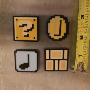 Puede incluir: Cuatro imanes pixelados con bordes negros. Los imanes incluyen un bloque amarillo con un signo de interrogaci&oacute;n, una moneda de oro, una nota musical blanca y un bloque de ladrillos marr&oacute;n. Los imanes miden aproximadamente 2,5 cm.