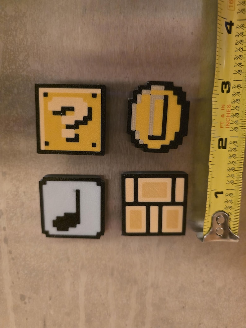 Puede incluir: Cuatro imanes pixelados con bordes negros. Los imanes incluyen un bloque amarillo con un signo de interrogaci&oacute;n, una moneda de oro, una nota musical blanca y un bloque de ladrillos marr&oacute;n. Los imanes miden aproximadamente 2,5 cm.
