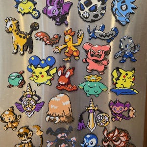 Peut inclure: Une collection de vingt-quatre aimants en pixel art fabriqués à partir de perles en plastique colorées. L'ensemble présente divers motifs de créatures stylisées dans des tons vifs de jaune, bleu, rose, violet, orange et marron, disposés en grille.
