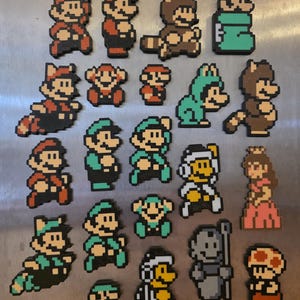 Puede incluir: Una colecci&oacute;n de imanes de pixel art con personajes de la serie de videojuegos Super Mario. Los imanes representan a Mario, Luigi, la Princesa Peach y otros personajes en varias poses y disfraces. Los imanes est&aacute;n hechos de cuentas de pl&aacute;stico de colores.