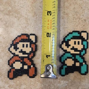 Puede incluir: Dos figuras de pixel art de personajes de videojuegos, una en rojo y otra en verde azulado, de aproximadamente 3,8 cm de altura cada una. Una cinta m&eacute;trica amarilla se coloca entre ellas, mostrando la altura de las figuras en pulgadas.
