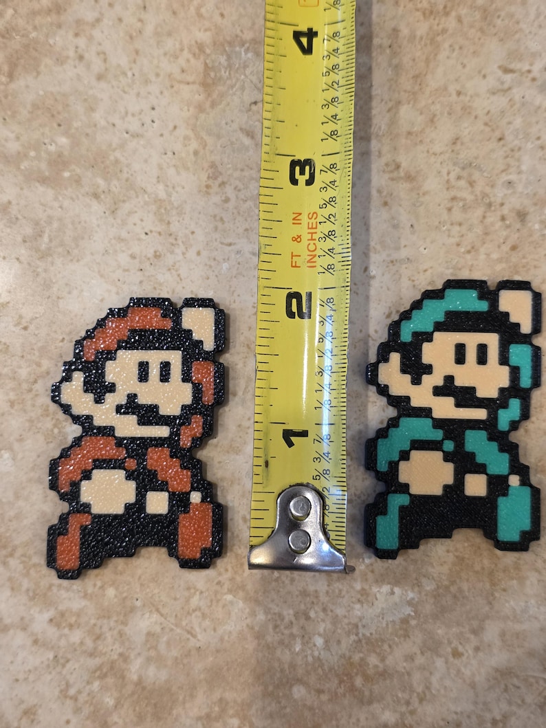 Puede incluir: Dos figuras de pixel art de personajes de videojuegos, una en rojo y otra en verde azulado, de aproximadamente 3,8 cm de altura cada una. Una cinta m&eacute;trica amarilla se coloca entre ellas, mostrando la altura de las figuras en pulgadas.