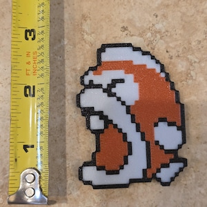 Puede incluir: Un personaje de videojuego pixelado, naranja, blanco y negro, de aproximadamente 7,6 cm de altura, se muestra junto a una cinta m&eacute;trica amarilla. El personaje tiene un contorno negro y est&aacute; sobre una superficie de tono neutro.