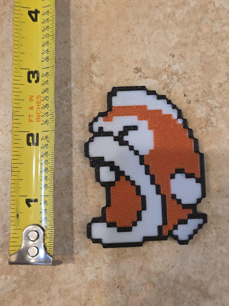 Puede incluir: Un personaje de videojuego pixelado, naranja, blanco y negro, de aproximadamente 7,6 cm de altura, se muestra junto a una cinta m&eacute;trica amarilla. El personaje tiene un contorno negro y est&aacute; sobre una superficie de tono neutro.