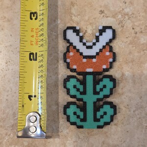 Puede incluir: Una planta Pira&ntilde;a pixelada, un personaje de la serie de videojuegos Super Mario. La planta es naranja con dientes blancos y un tallo y hojas verdes. La planta mide aproximadamente 6,35 cm de altura.