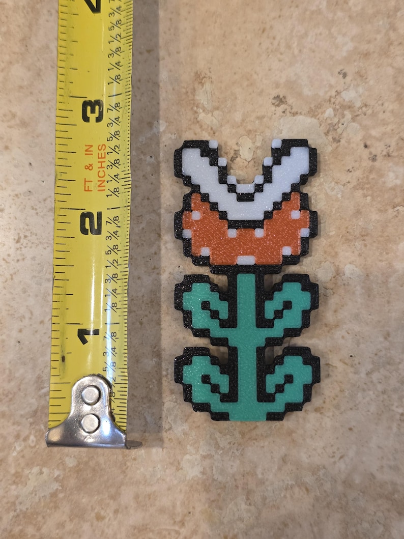 Puede incluir: Una planta Pira&ntilde;a pixelada, un personaje de la serie de videojuegos Super Mario. La planta es naranja con dientes blancos y un tallo y hojas verdes. La planta mide aproximadamente 6,35 cm de altura.