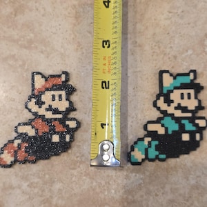 Puede incluir: Dos figuras de pixel art de un personaje de videojuego, una en marr&oacute;n y naranja, la otra en verde azulado y azul. Las figuras miden aproximadamente 5 cm de largo. Una cinta m&eacute;trica amarilla est&aacute; en el fondo.