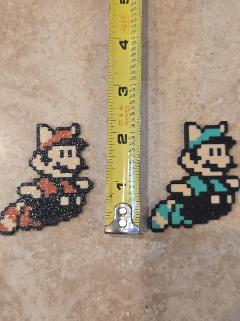 Puede incluir: Dos figuras de pixel art de un personaje de videojuego, una en marr&oacute;n y naranja, la otra en verde azulado y azul. Las figuras miden aproximadamente 5 cm de largo. Una cinta m&eacute;trica amarilla est&aacute; en el fondo.
