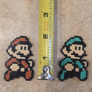 Puede incluir: Figuras de arte pixelado de Mario y Luigi, cada una de aproximadamente 5 cm de altura. Mario es rojo y Luigi verde, ambos con contornos negros y tonos de piel beige. Una cinta m&eacute;trica amarilla est&aacute; entre ellos.