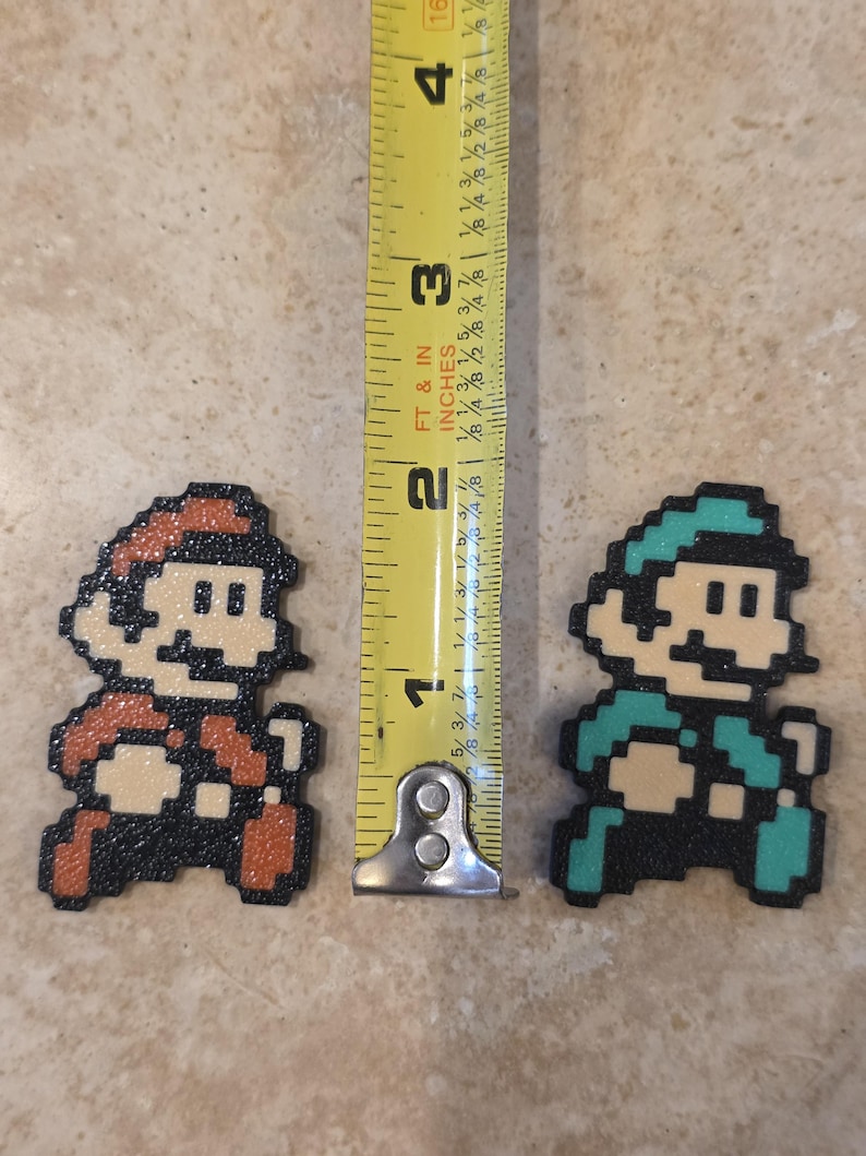 Puede incluir: Figuras de arte pixelado de Mario y Luigi, cada una de aproximadamente 5 cm de altura. Mario es rojo y Luigi verde, ambos con contornos negros y tonos de piel beige. Una cinta m&eacute;trica amarilla est&aacute; entre ellos.