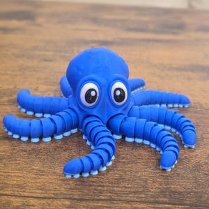 May include: A blue articulated octopus figurine with a rounded head and eight segmented, flexible tentacles. The creature features large, white, oval-shaped eyes with black pupils and small, light blue circular suction cups on the underside of each limb.