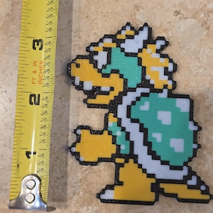 Puede incluir: Una figura pixelada de Bowser, de aproximadamente 7,6 cm de altura, con caparaz&oacute;n verde, vientre y cabeza amarillos y detalles blancos. La figura tiene un acabado brillante y est&aacute; delineada en negro. Una cinta m&eacute;trica amarilla est&aacute; en la imagen.